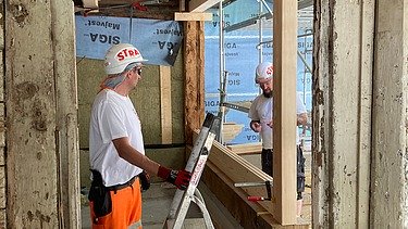 Zwei STRABAG Zimmermänner mit Helmen und Schutzkleidung arbeiten an einer Holzkonstruktion in einem offenen Raum.