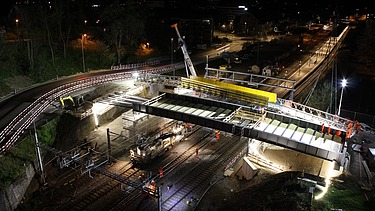 Eine Baustelle wird nachts unter hellem Arbeitslicht beleuchtet. Arbeiter und Maschinen montieren ein grosses Brückenelement über einer Bahnlinie.