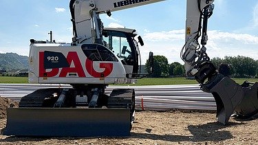 Ein weisser STRABAG Liebherr-Raupenbagger steht einsatzbereit auf einer Erd- und Kiesfläche vor einem Baucontainer.