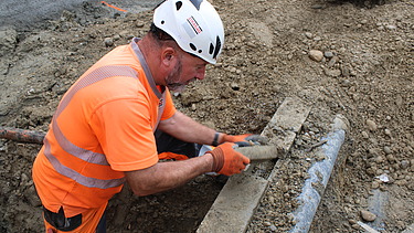 Ein Bauarbeiter mit orangefarbener Arbeitskleidung und weissem Helm legt ein eine alte Werkleitung frei 