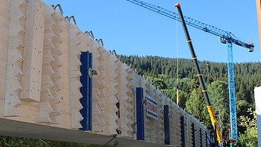 Ein langer Holzträger mit STRABAG Logo liegt auf einer Transportvorrichtung im Hintergrund heben zwei Kräne den Träger vor bewaldeten Hügeln an.