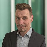 Kaufmännischer Bereichsleiter im Anzug lächelt in die Kamera. Im Hintergrund ist ein Lift zu sehen.