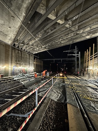 Nächtliche Baustelle unter einer Betonbrücke mit beleuchteten Gleisen und Baugeräten.