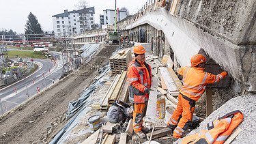 Zwei Bauarbeiter in leuchtenden Jacken inspizieren eine Bohrpfahlwand auf einer STRABAG Baustelle in Küssnacht. Im Hintergrund sind Absperrungen und ein Kran.