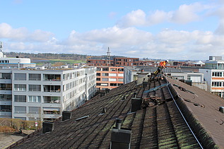 STRABAG Sicherheitsbeauftragter auf einem Dach mit Sicherungsseil. Im Hintergrund weitere Wohnblocks und eine blauer Himmer.