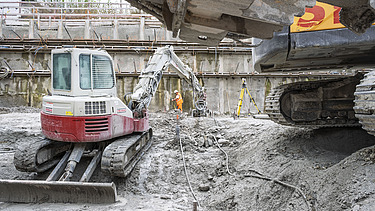 Bild aus der Baugrube im Vordergrund ein Minibagger im Hintergrund ein grosser Bagger bei dem die Schaufel ins Bild rein ragt 