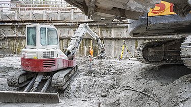 Bild aus der Baugrube im Vordergrund ein Minibagger im Hintergrund ein grosser Bagger bei dem die Schaufel ins Bild rein ragt 