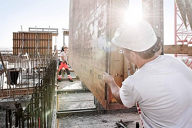 Ein Bauarbeiter richtet eine Schalwand auf einer Hochbaustelle aus. Die Sonne scheint durch die Baustelle, im Hintergrund sind weitere Schalungen zu sehen.