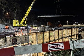 Im Vordergrund STRABAG Absperr-Latten im Hintergrund ein Kran der ein  24 Tonnen Stahlträger hebt