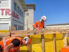 Zwei Lernende in orangefarbener STRABAG Arbeitskleidung arbeiten an einer gelben Verschalung auf einer Baustelle.