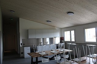 Heller Küchen- und Essraum mit weissen Schränken, grossen Tischen und gestapelten Stühlen.