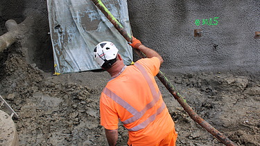 Ein Bauarbeiter mit orangefarbener Arbeitskleidung und weissem Helm hält ein langes Rohr in dem Graben an der Baustelle.