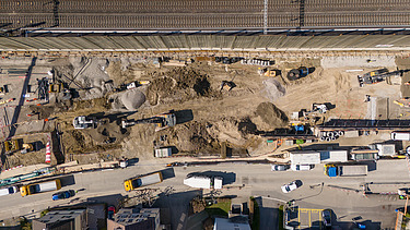 Drohnenaufnahme der Baugrube von oben, zu sehen sind mehrere Bagger, die Bahnlinie welche parallel zur Baugrube verläuft sowie Baustellenfahrzeuge