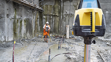 Ein Bauarbeiter ist in der Baugrube beim Vermessen mit Laser, im Vordergrund das Lasergerät im Hintergrund er