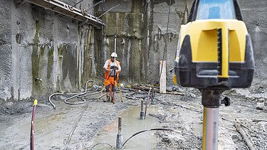 Ein Bauarbeiter ist in der Baugrube beim Vermessen mit Laser, im Vordergrund das Lasergerät im Hintergrund er