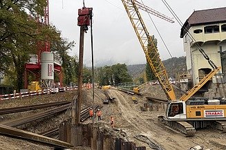 Luftbild: Baustelle am Fluss mit grossem rotem Kran, gelbem Bagger, Spundwänden und Baugrube neben einer Staumauer im Herbst.