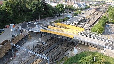 Fertig eingesetzte Bahnbrücke in Küssnacht. Die Brücke führt über mehrere Bahngleise. Im Hintergrund sind Strassen, Gebäude und ein Bahnhof zu erkennen.