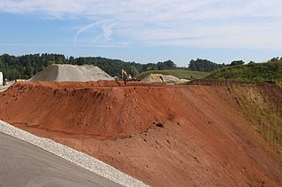 Erddeponie mit grossen Aushubmengen im Vordergrund eine rötliche Böschung, im Hintergrund sind Maschinen und Ablagerungen zu sehen.