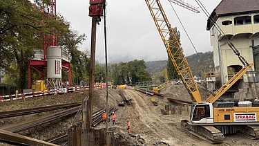 Baustelle am Flussufer: Sicht vom Wasser auf eine Staumauer, roten Kran und Spundwände der Baugrube.