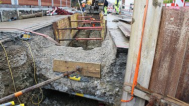 Baustelle an der Amriswilerstrasse - Tiefbau STRABAG Offene Baugrube mit Stahlstützen zur Sicherung der Seitenwände. Im Vordergrund verlaufen Kabel und Rohre entlang der Grube, während im Hintergrund Bauarbeiter und ein Bagger bei der Arbeit. Eine Schalung stabilisiert die Grabenwände, Sicherheitsabsperrungen und Markierungen kennzeichnen den Baustellenbereich.