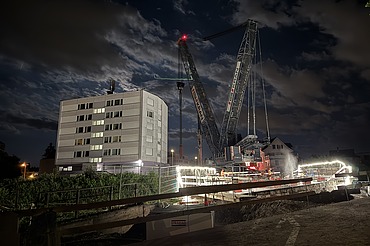 Nachtaufnahme einer STRABAG-Baustelle. Ein grosser Kran steht beleuchtet neben einem Wohnhaus unter dem klaren Nachthimmel.