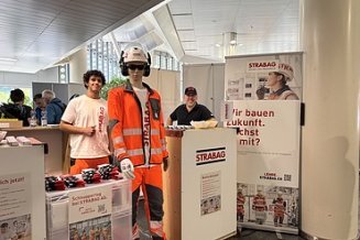 Messestand der STRABAG AG mit zwei Mitarbeitenden und einer Schaufensterpuppe in oranger STRABAG-Arbeitskleidung mit Helm.