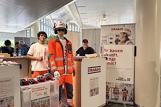 Messestand der STRABAG AG mit zwei Mitarbeitenden und einer Schaufensterpuppe in oranger STRABAG-Arbeitskleidung mit Helm.