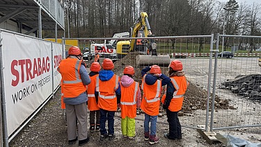 Eine Gruppe von Schulkindern in orangefarbenen Schutzwesten und Helmen steht vor einem Zaun und beobachtet einen Bagger bei Bauarbeiten auf einer Baustelle in Kloten.