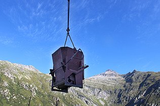 Ein Kübel für Beton wird vor dem blauen Himmel und einer Bergkulisse in der Schweiz über die STRABAG Baustelle gehoben.