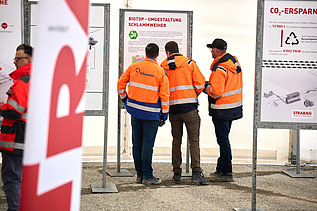 Mehrere Personen in orangefarbener Schutzkleidung stehen vor Informationstafeln einer Baustelle. Im Vordergrund ist unscharf eine STRABAG-Fahne zu sehen.