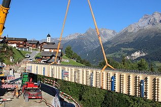 Ein grosser Kran hebt einen langen Holzträger an, während dieser langsam über die Baustelle und vor der Bergkulisse schwebt.
