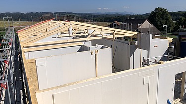 Rohbau der Labor-Erweiterung mit weissen vorgefertigten Wänden und sichtbaren Holzbalken für das Dach, eingerahmt von Baugerüsten, im Hintergrund ländliche Umgebung mit Feldern, Bäumen und Hügeln.