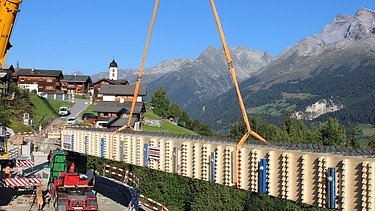 Ein grosser Kran hebt einen langen Holzträger an, während dieser langsam über die Baustelle und vor der Bergkulisse schwebt.