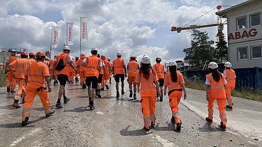 Eine Gruppe von STRABAG-Lernende in orangefarbener Arbeitskleidung und weissen Helmen geht auf eine Baustelle.