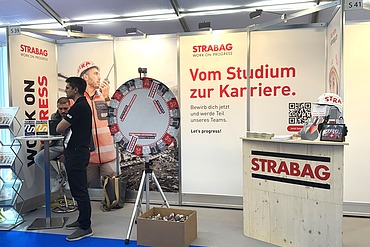 Mann steht vor dem STRABAG-Stand an der KTH 2026. Man sieht links den STRABAG Holzmessestand, im Hintergrund eine Messewand mit verschiedenen Bildern. Im rechten Teil des Bildes stehen zwei Männer an einem Bartisch. 