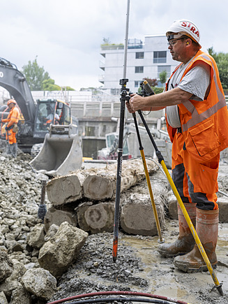 Der Polier misst mit dem Vermessungsgerät in der Baugrube. Im Hintergrund ein grosser Bagger beim Aushub. 