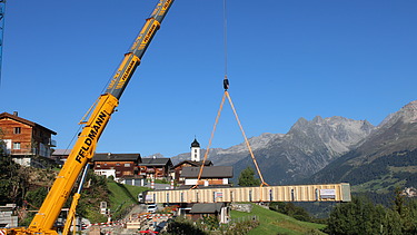 Ein Pneukran hebt einen langen Holzträger über ein Bergdorf im Vordergrund ein Kranführer und Monteure, im Hintergrund sind die Alpen zu sehen.