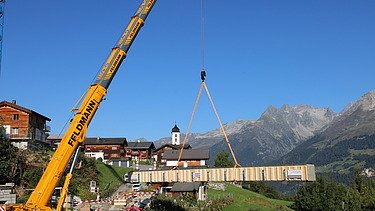 Ein Pneukran hebt einen langen Holzträger über ein Bergdorf im Vordergrund ein Kranführer und Monteure, im Hintergrund sind die Alpen zu sehen.