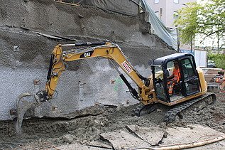 Ein gelber Bagger mit STRABAG-Logo arbeitet in einem Graben mit leichter Hanglage, ein Bauarbeiter sitzt im Bagger.