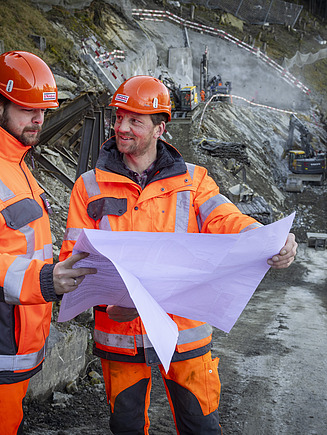 Zwei STRABAG Mitarbeiter in Arbeitsklamotten lesen einen Bauplan im Hintergrund sieht man den Sisikoner Tunnel und die Baugrube.