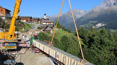 Ein großer Pneukran hebt einen langen Holzträger langsam in die Höhe; darunter stehen Arbeiter und im Hintergrund sieht man ein Bergdorf vor einer beeindruckenden Alpenkulisse.