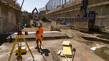 Rüschlikon Bahnhof Süd – STRABAG Tiefbau und Spezialtiefbau Vermesser steht in der Baugrube am Ausmessen. Rechts daneben hebt ein Bagger gerade ein Loch aus.