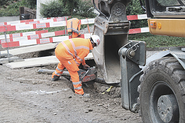 Zwei STRABAG - Mitarbeiter arbeiten an einer geöffneten Leitungstrasse. Sie überprüfen oder verlegen Rohre im Boden, während ein Bagger daneben steht. Absperrungen sichern die Baustelle auf einer Strasse.