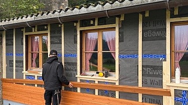 Blick auf die Holzfassade des Pfadiheims an der gerade ein Lernender dabei ist die neuen Holz Latten für die Fassade zusammenzustecken