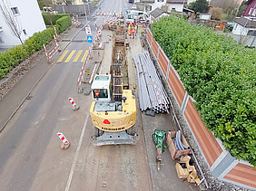 Luftaufnahme einer Baustelle mit offener Baugrube und einem Bagger. Die Baugrube ist mit Absperrungen und Verkehrsschildern gesichert, während Werkleitungen und Baumaterialien entlang der Strasse gestapelt sind. Im Hintergrund sind Bauarbeiter bei der Installation und Verlegung von Leitungen tätig. Die Baustelle befindet sich an einer Strasse mit markierten Fussgängerüberwegen und einer geregelten Verkehrsführung. 