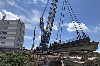 Ein 750-Tonnen-Fangkran der STRABAG hebt ein Brückenelement aus der Baustelle; mehrere Kräne und Mitarbeitende sichern den Bereich.
