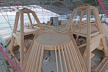 Eine grosse Holzkonstruktion im Umbau unter einer Schutzhalle. Zu sehen sind Balken, Träger und Stützen aus Holz, an denen Zimmerleute arbeiten. Das Bauwerk befindet sich auf einer Baustelle am Bahnhofquai.