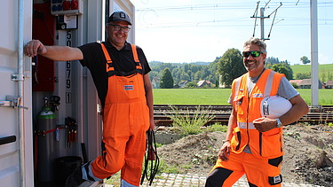 Zwei Männer in orangefarbener Bauausrüstung stehen lachend vor einem geöffneten Container auf der Baustelle.