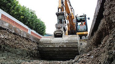 Bagger beim Aushub einer Baugrube, aufgenommen aus der Perspektive der Grubensohle. Der Baggerführer sitzt in der Kabine und steuert die Schaufel, die gerade Erdmaterial bewegt.