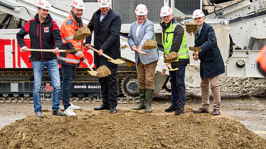 Mehrere Personen in Schutzhelmen und Arbeitskleidung führen gemeinsam einen Spatenstich auf einer Baustelle durch. Hinter ihnen steht schweres Baugerät.